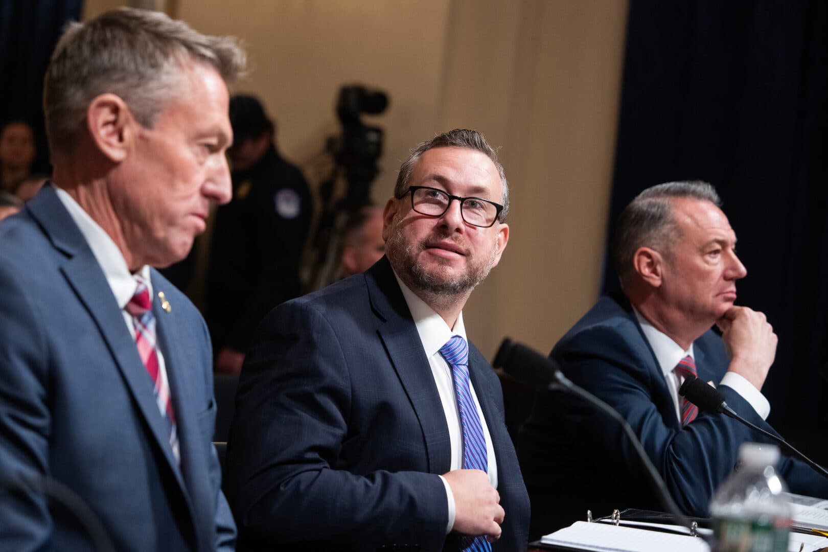 USCIS Director Joseph Edlow seated at a hearing table, flanked by two other officials in suits.